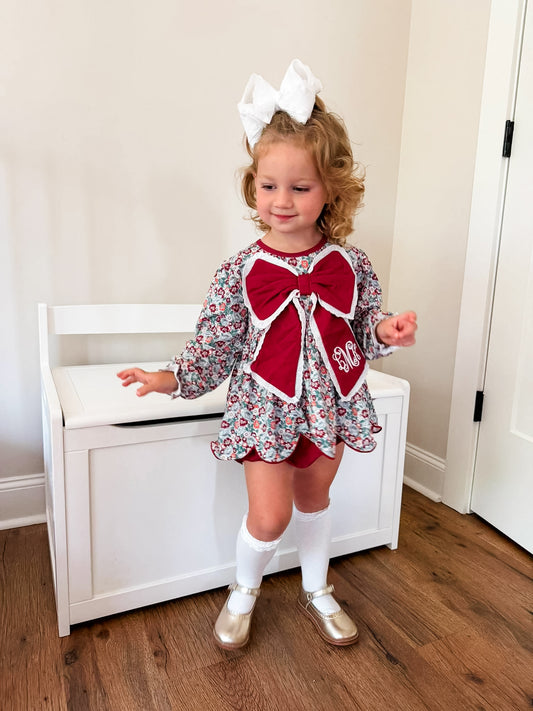 Child wearing a festive outfit with a large red bow and white shoes, standing in a room.